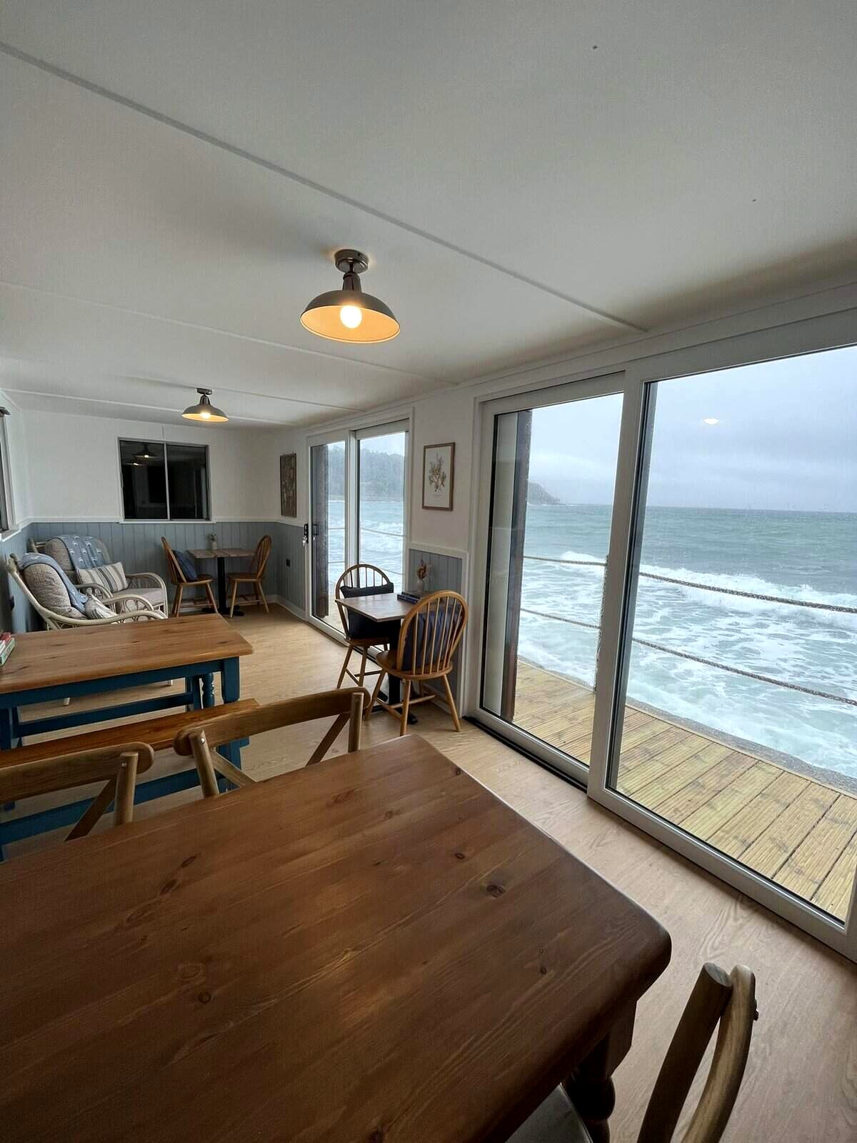 Inside the café: wooden tables and chairs, a blue-panelled wall and floor-to-ceiling glass doors onto a deck, with waves breaking just outside
