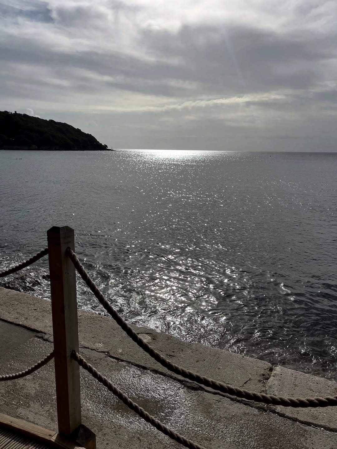 View from the café's edge: a rope rail on the sea wall, silver light on the water of Falmouth Bay and a wooded headland