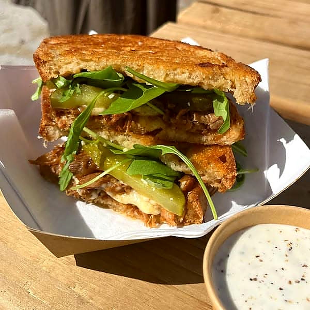 A toastie on golden toasted bread, filled with rocket and pickles, in a paper tray with a pot of dip
